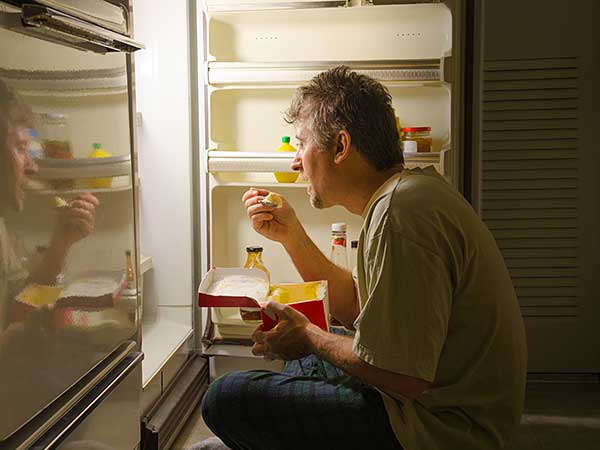 Man with sleep eating disorder eating at night in front of refrigerator.